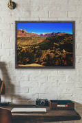landscape art print depicting a desert canyon with red rock cliffs and a blue sky, displayed on a white brick wall above a wooden desk with a camera and laptop.