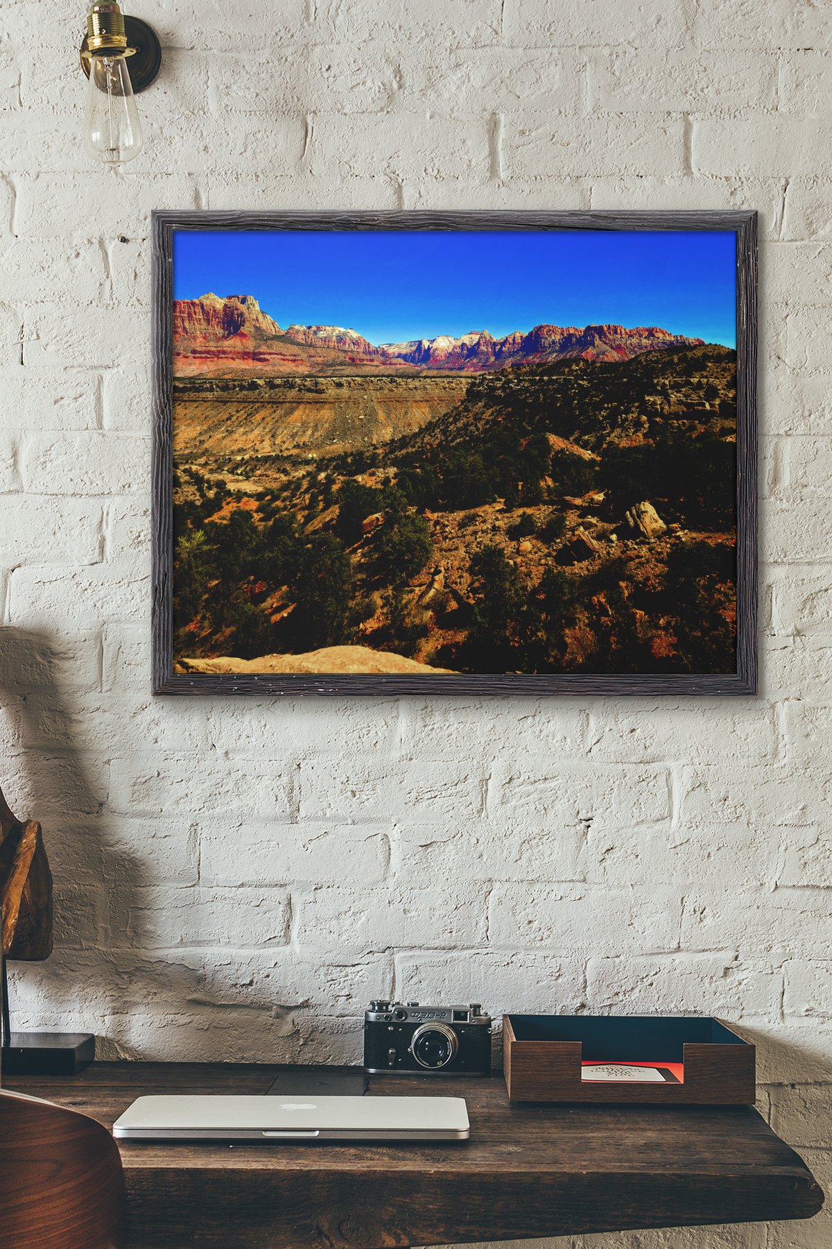 landscape art print depicting a desert canyon with red rock cliffs and a blue sky, displayed on a white brick wall above a wooden desk with a camera and laptop.